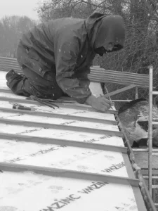Couvreur agenouillé sur un toit en bacs métalliques sous une chute de neige, traçant une ligne au crayon.