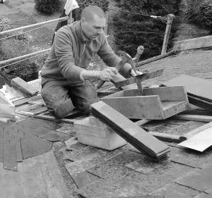 Un couvreur à genoux ajuste et fixe des planches de bois pour former le coffrage d'une rive de toit.